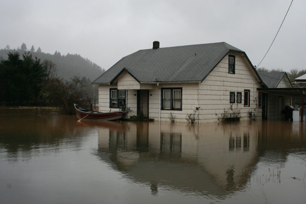 An older tan home in flooded water with a drift boat floating in front of it.