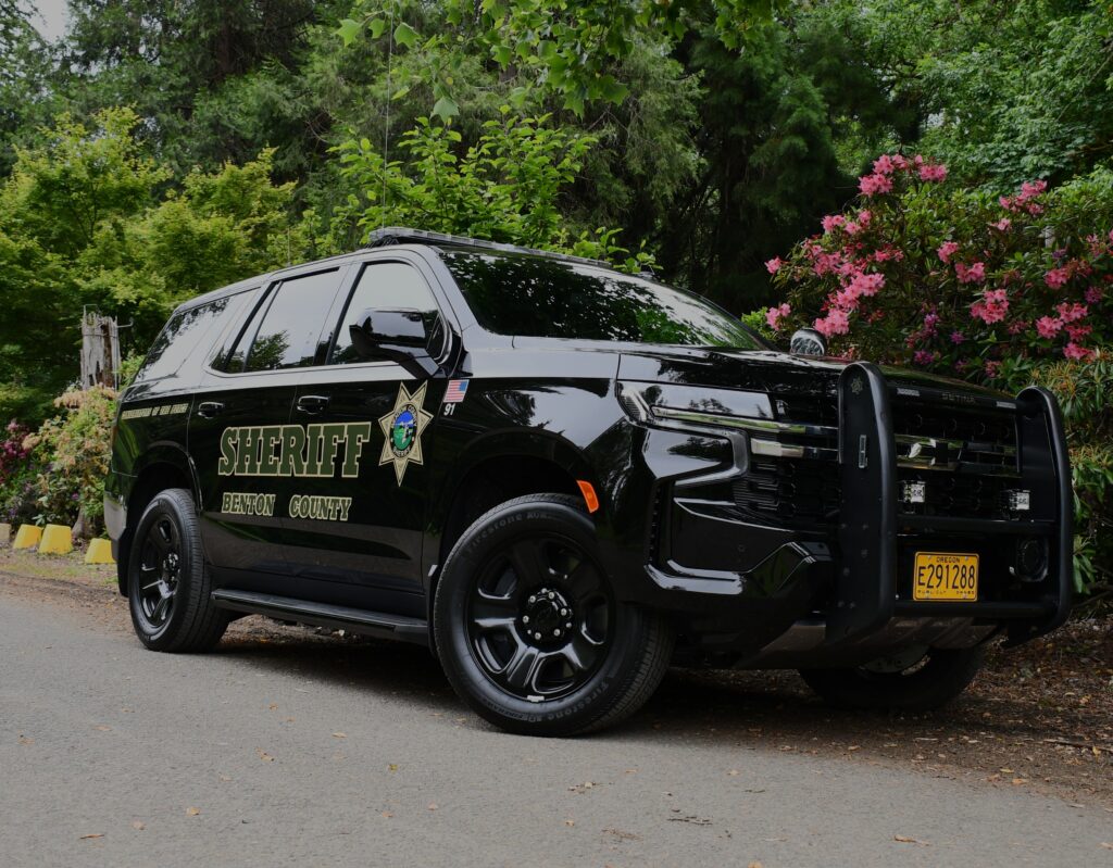 A dark colored patrol SUV with lights on, 7-point gold star, and the words SHERIFF BENTON COUNTY on the side with trees in the background.